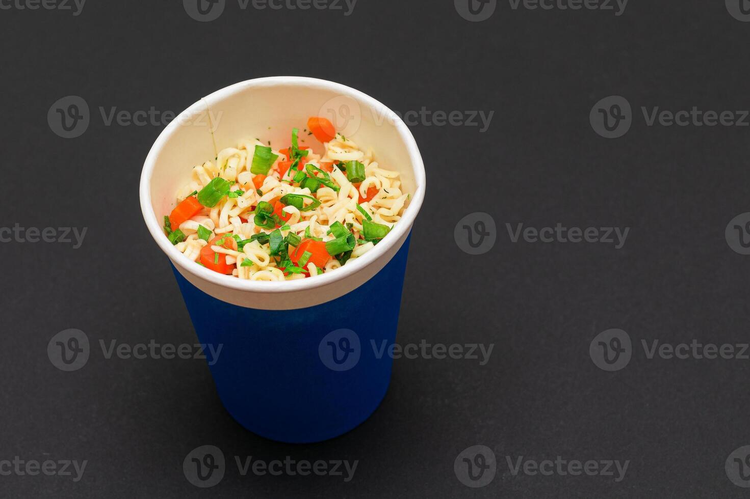 Prepared Instant Noodles with Carrot and Greens in Blue Cup on Black Background. Quick Lunch or Unhealthy Fast Food photo