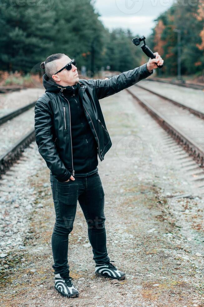 Blogger in Sunglasses Making Selfie or Streaming at the Pine Forest Using Action Camera with Gimbal Camera Stabilizer. Handsome Man in Black Clothes Making Photo