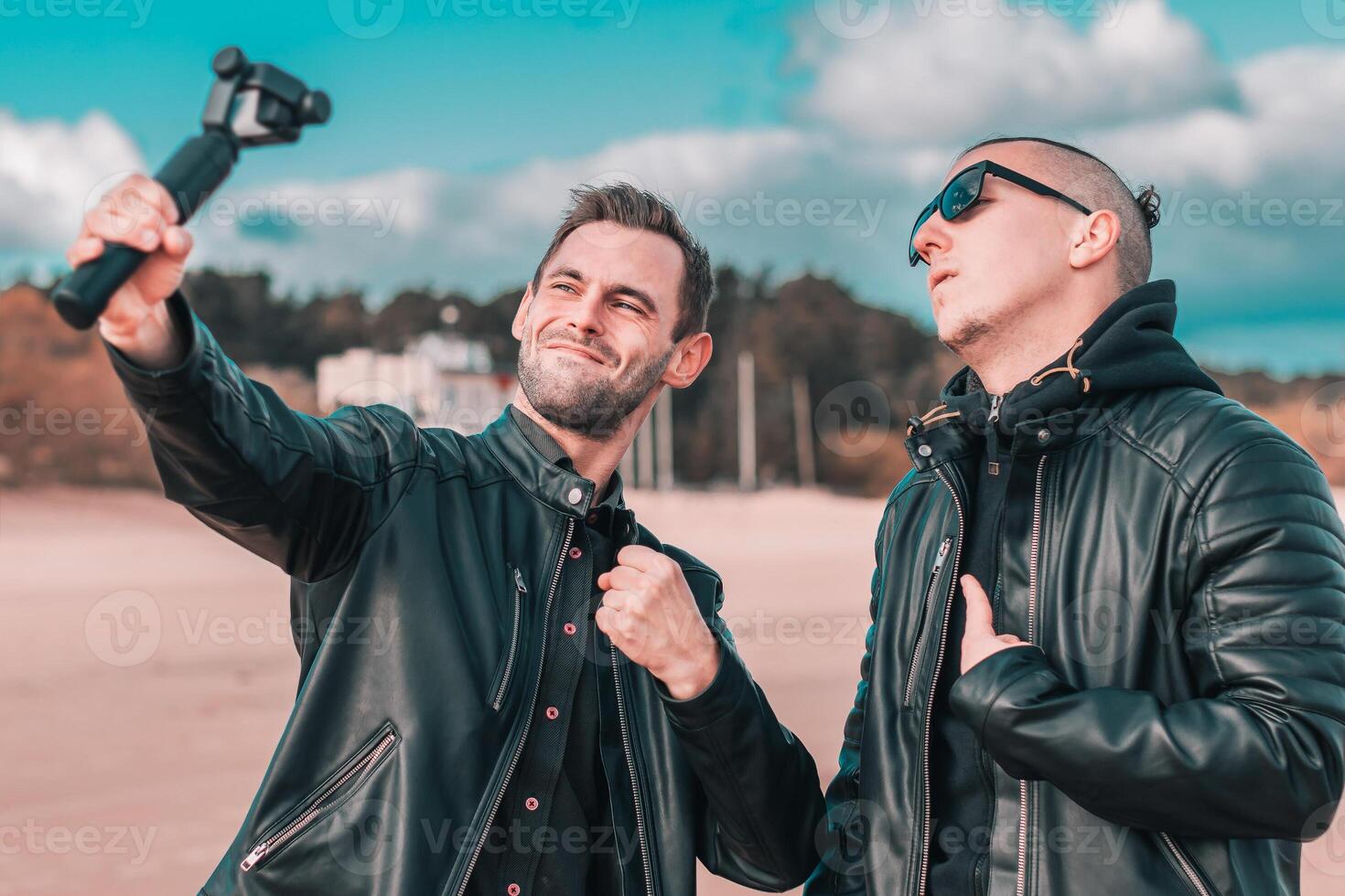 Two Handsome Male Friends Making Selfie Using Action Camera with Gimbal Stabilizer at the Beach. Youthful Men in Black Clothes Having Fun by Making Photos