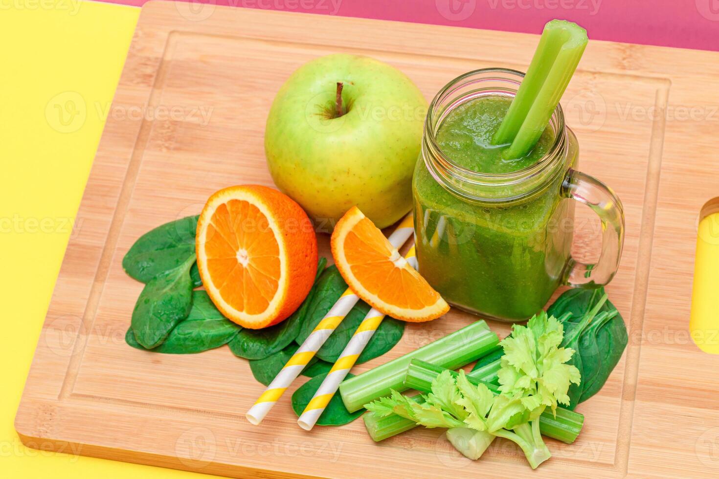 Fresh Green Smoothie of Apple, Celery, Spinach and Orange in Glass Smoothie Jar with Yellow Cocktail Straw on Wooden Cutting Board. Vegan Detox Drink. Vegetarian Culture. Healthy Eating and Fruit Diet photo