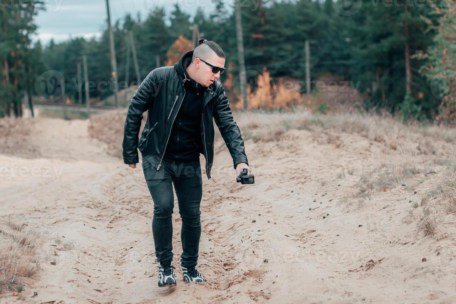 Youthful Blogger in Black Clothes and Sunglasses Making Using Action Camera with Gimbal Camera Stabilizer in Pine Forest photo