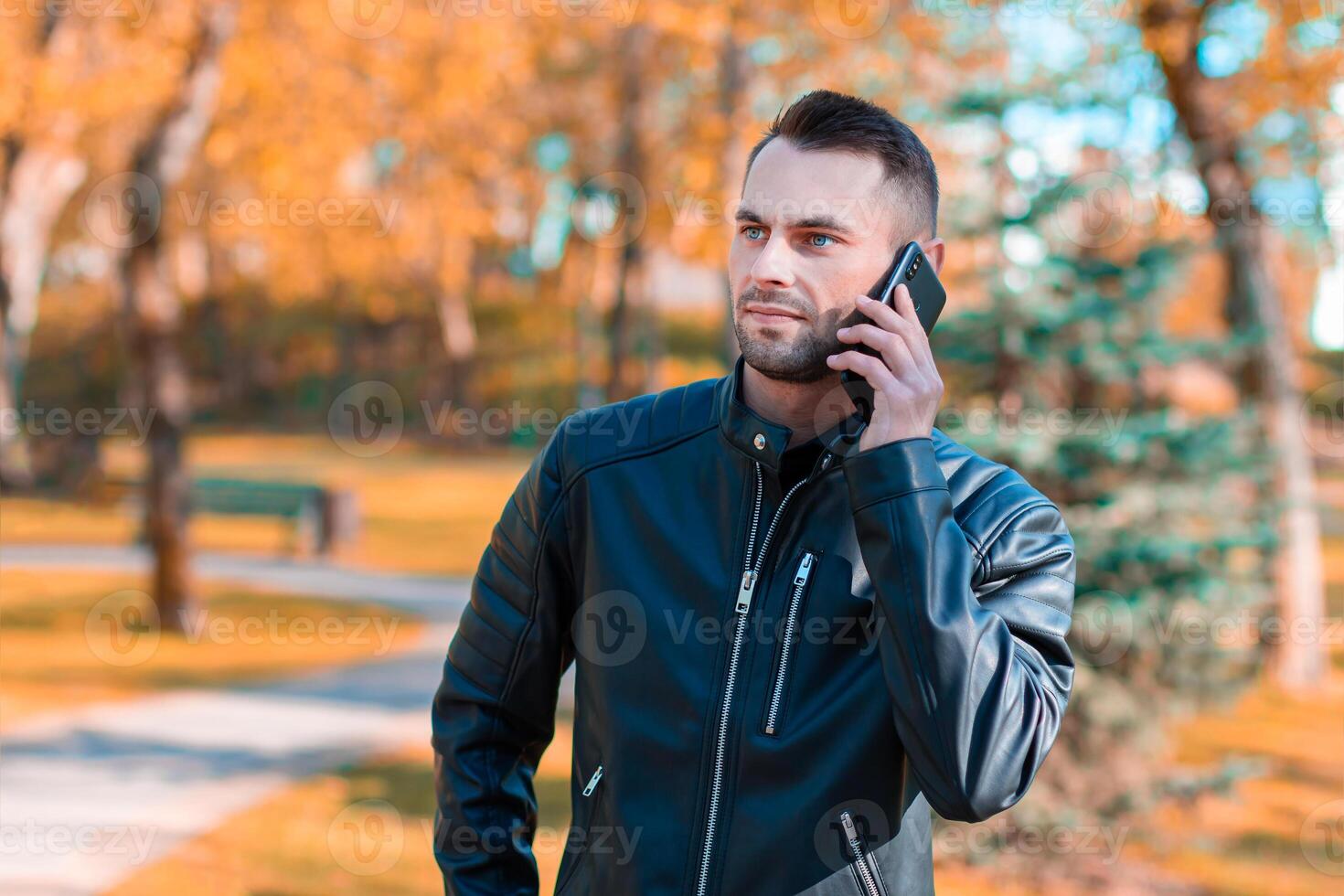 Handsome Young Man Talking on the Phone at the Beautiful Autumn Park. Youthful Guy Using Smartphone for the Call Outdoors at Sunny Day photo