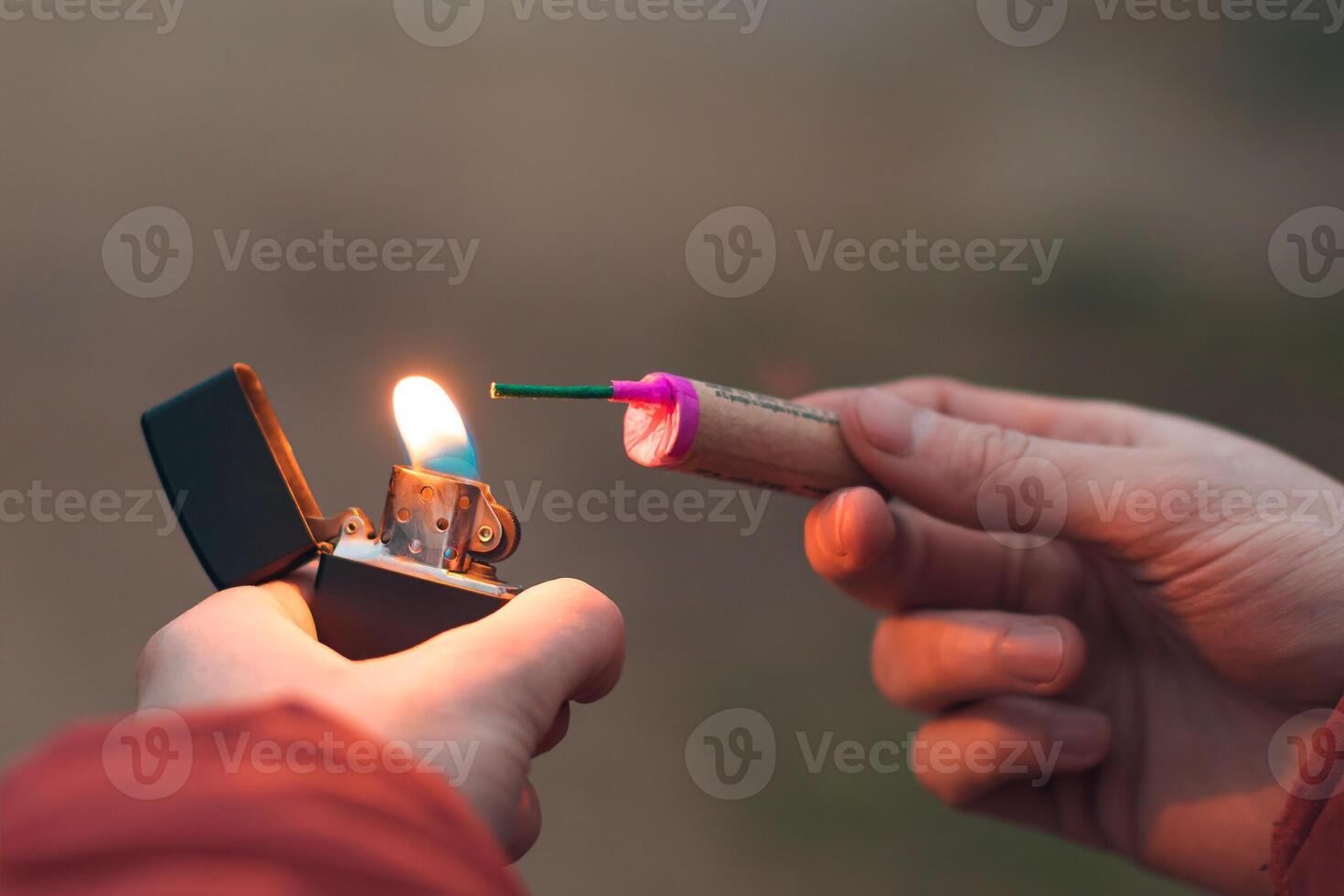 Young Man Lighting Up Firecracker in his Hand Using Gasoline Lighter. Guy Getting Ready for New Year Fun with Fireworks or Pyrotechnic Products - CloseUp Shot, Rear View photo