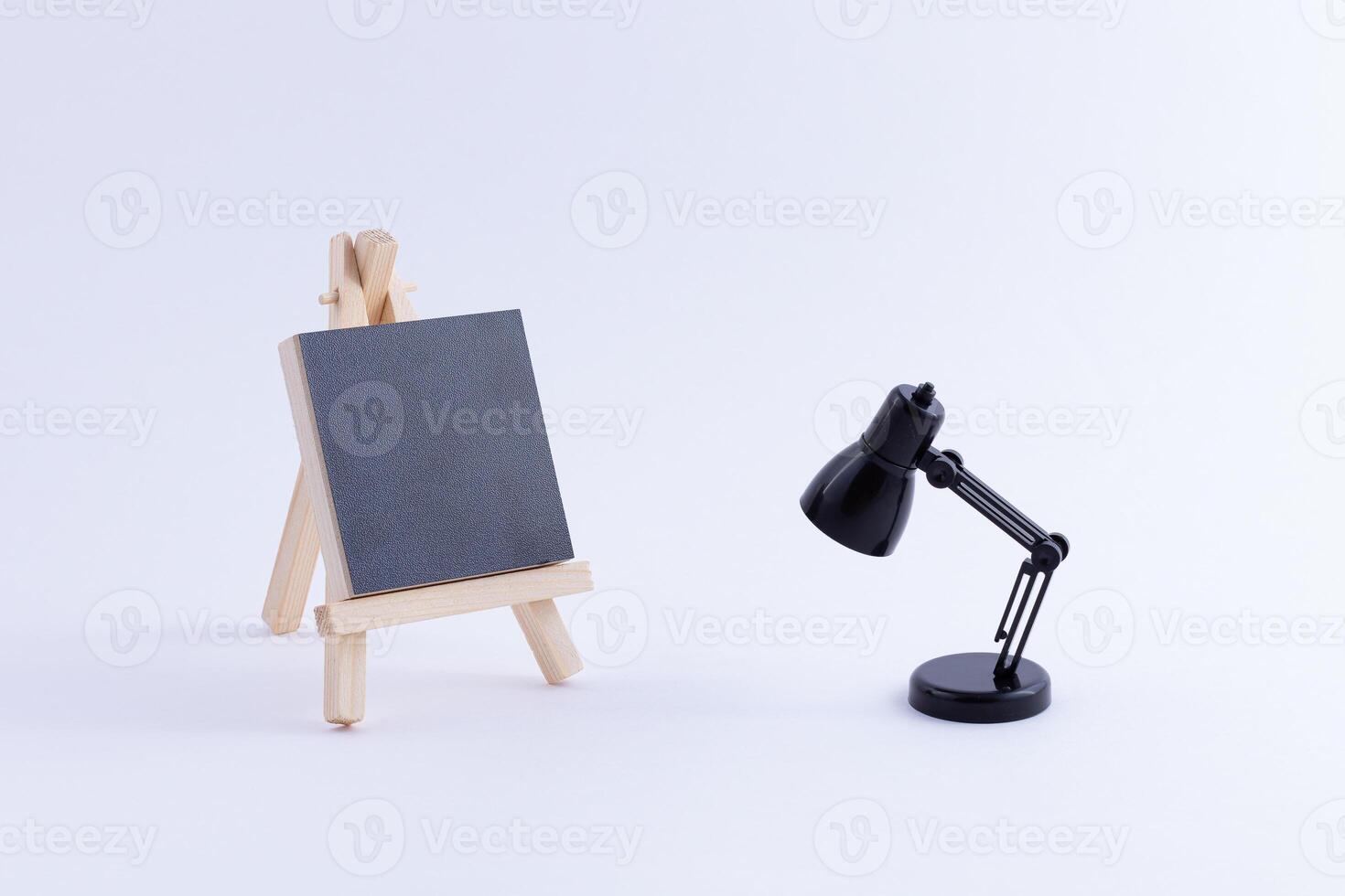 Wooden Easel Miniature with Blank Black Square Canvas for Artists and Painters - Mockup. Mini Wooden Stand with Clean Artboard and Small Black Table Lamp on White Background, Copy Space photo