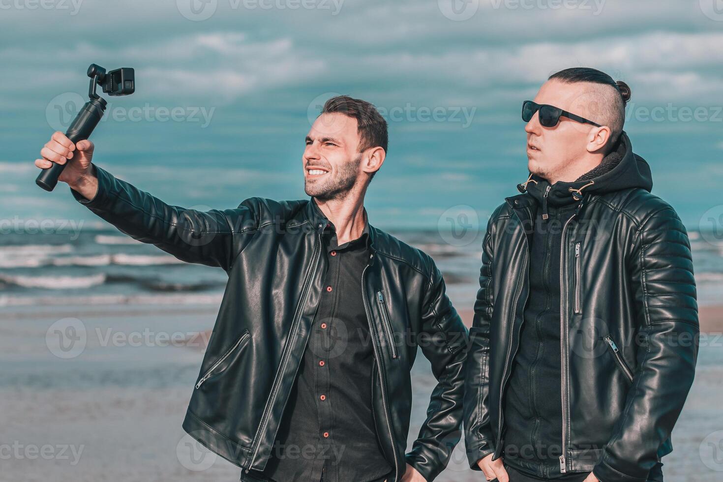 Two Handsome Smiling Friends Making Selfie Using Action Camera with Gimbal Stabilizer at the Beach. Youthful Men in Black Clothes Having Fun by Making Photos
