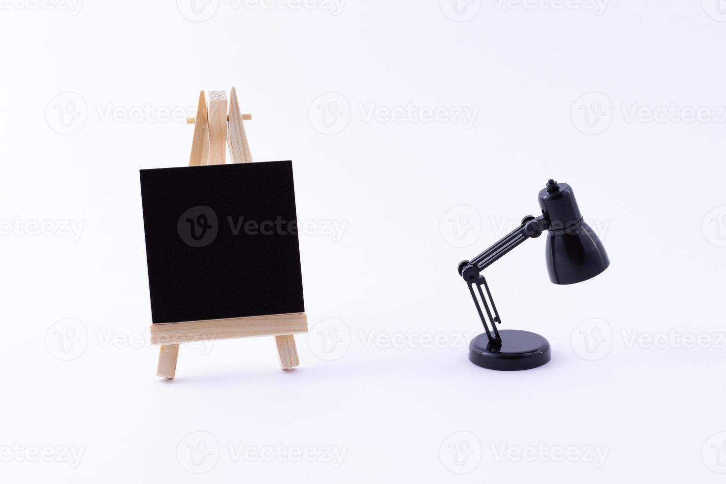 Wooden Easel Miniature with Blank Black Square Canvas for Artists and Painters - Mockup. Mini Wooden Stand with Clean Artboard and Small Black Table Lamp on White Background, Copy Space photo