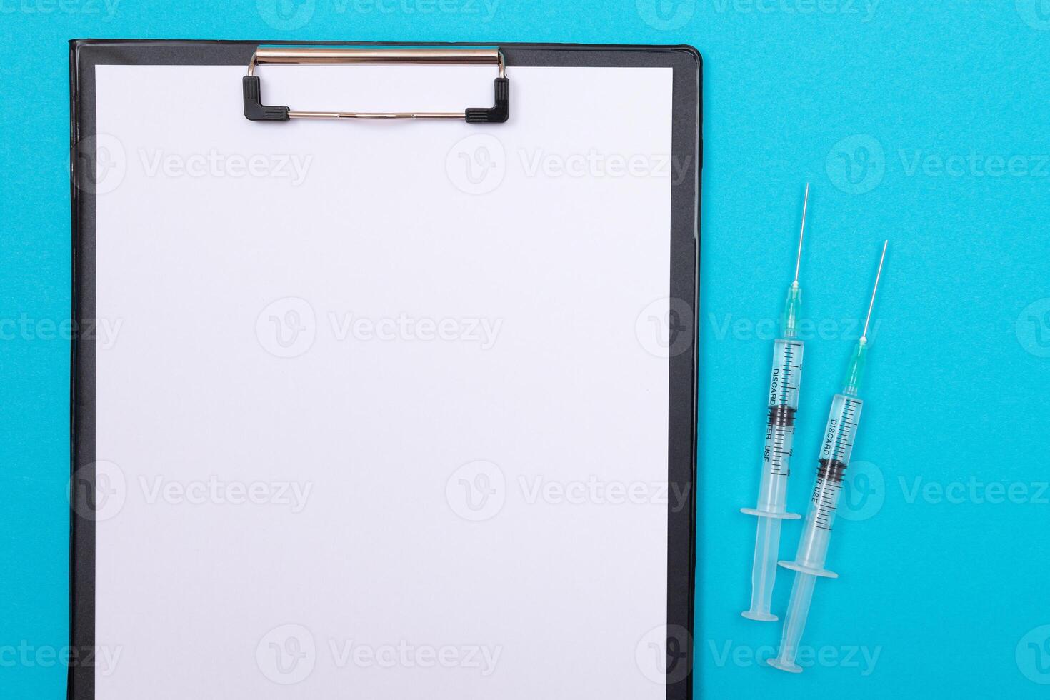 Vaccination, Immunology or Revaccination Concept - Two Medical Syringe Lying on Blue Table in Doctor's Office in a Hospital or Clinic. Black Clipboard with Sheet of Paper - Mock Up with Copy Space photo