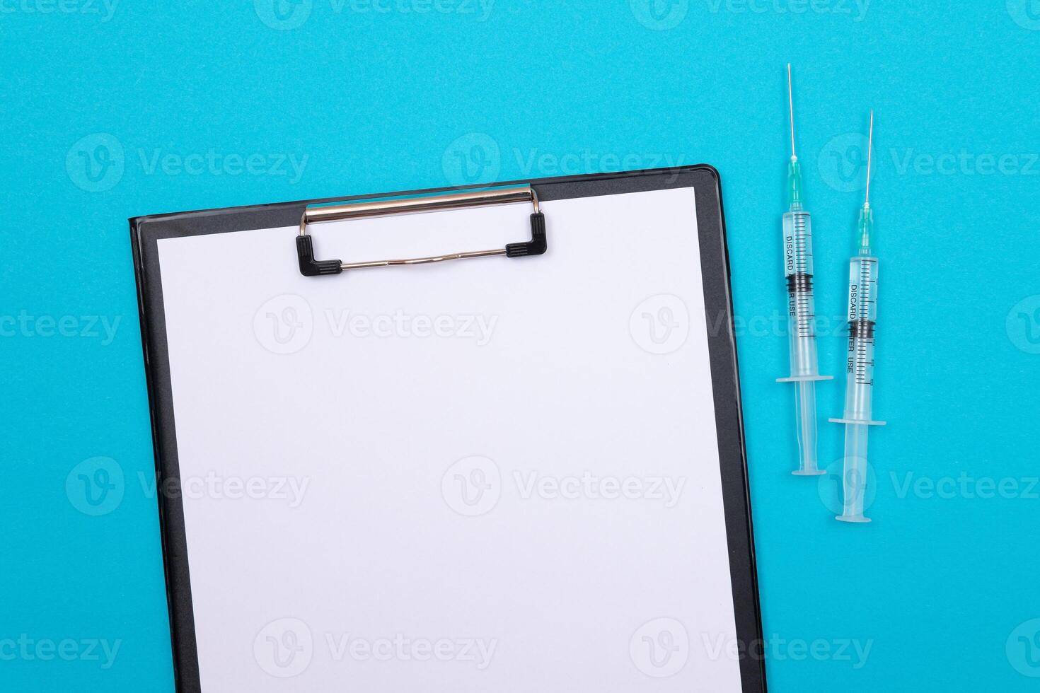 Vaccination, Immunology or Revaccination Concept - Two Medical Syringe Lying on Blue Table in Doctor's Office in a Hospital or Clinic. Black Clipboard with Sheet of Paper - Mock Up with Copy Space photo