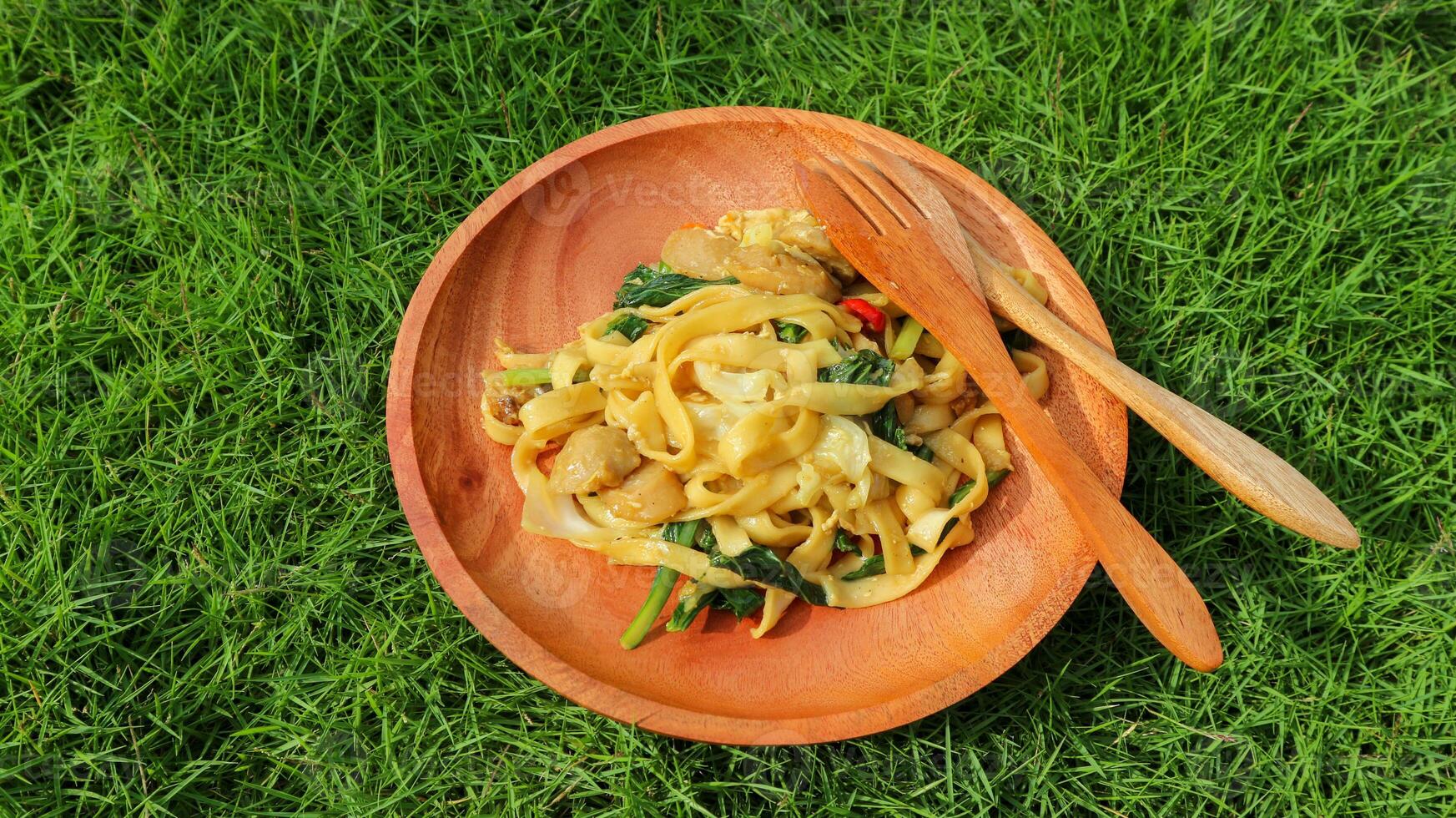 Kwetiaw or Kwetiau is Chinese food which is popular in Indonesia, cooked by frying added with chicken, seafood and complementary vegetables. Served in wooden plate on green grass background. photo