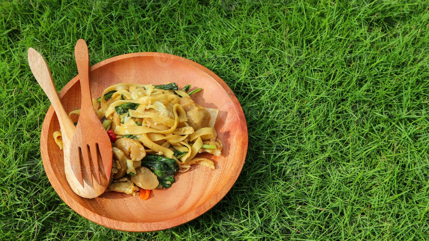 Kwetiaw or Kwetiau is Chinese food which is popular in Indonesia, cooked by frying added with chicken, seafood and complementary vegetables. Served in wooden plate on green grass background. photo