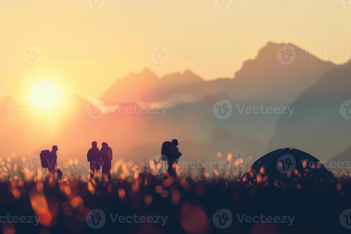 Sunset camping trip with silhouettes of hikers and tent in a meadow. Blurred mountains on a background. photo