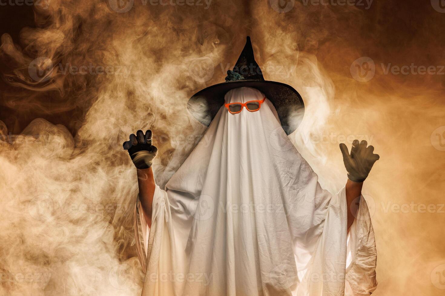 Man in scary ghost costume for Halloween and witch hat on orange mystical background in smoke photo