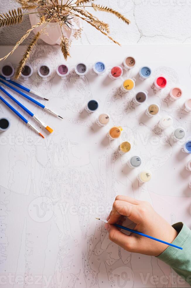 A girl paints a picture by numbers with a brush using a paint set top and vertical view photo