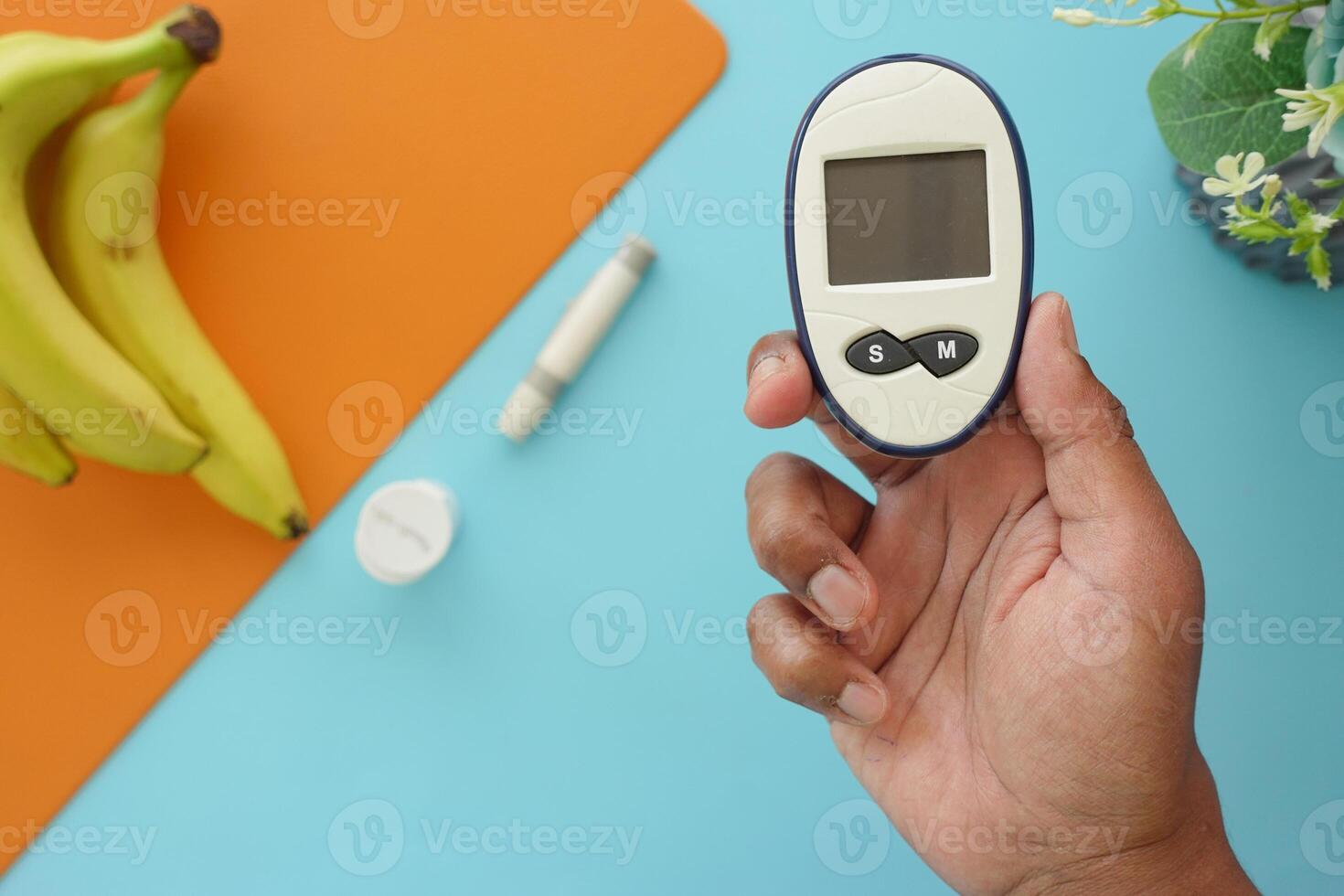 A hand is holding a glucometer alongside bananas and various essential diabetes supplies photo