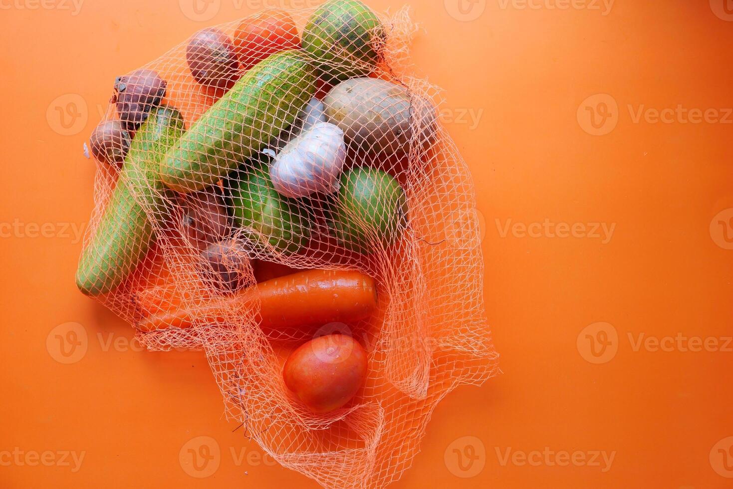 A variety of Fresh Vegetables presented in a Net Bag against a Vibrant Orange Background photo