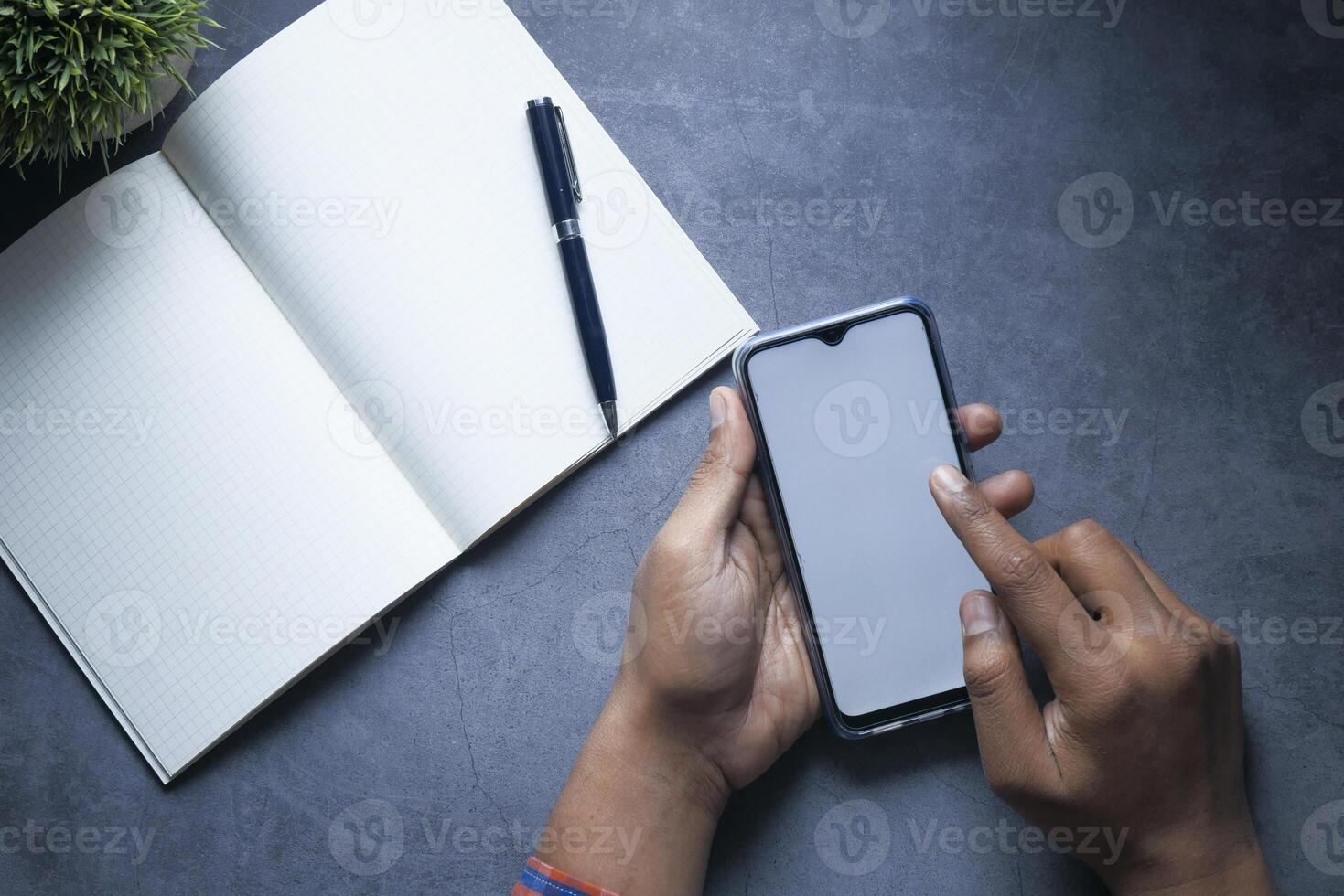 A person is using a smartphone while sitting next to a notebook and a pen on a dark surface photo