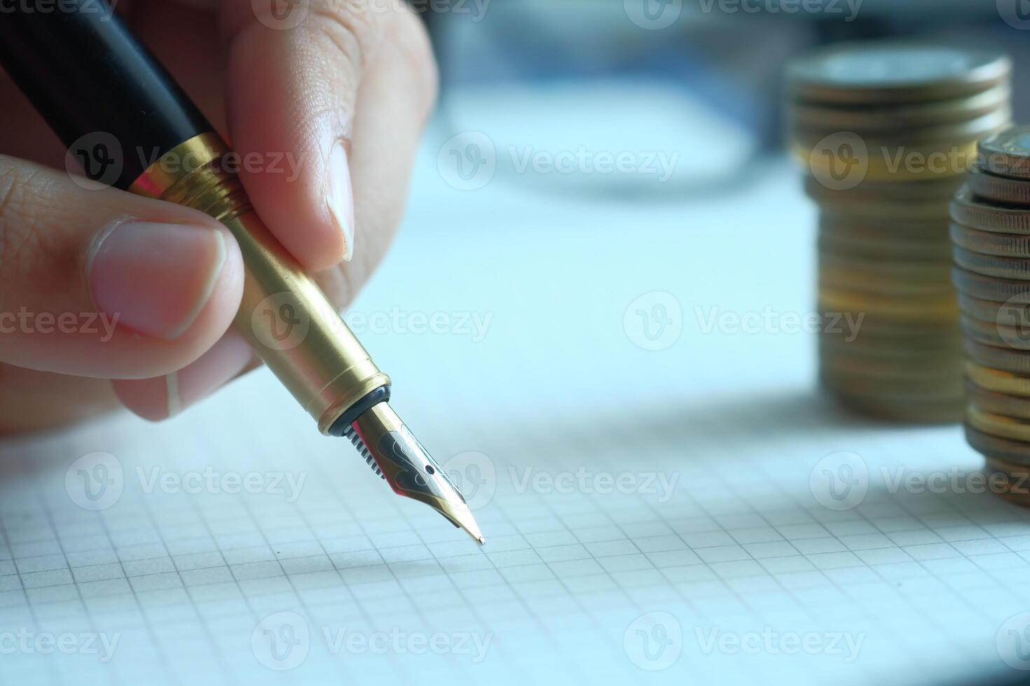 A closeup of a hand holding a fountain pen above a notepad with stacks of coins behind photo