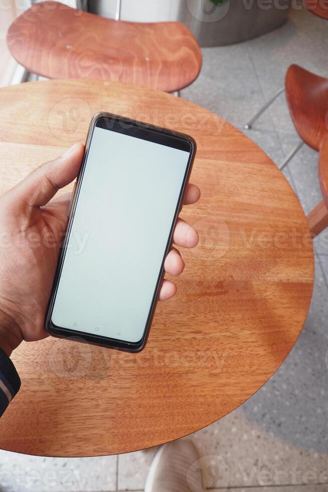 A hand is casually holding a smartphone while resting on a wooden tables surface photo