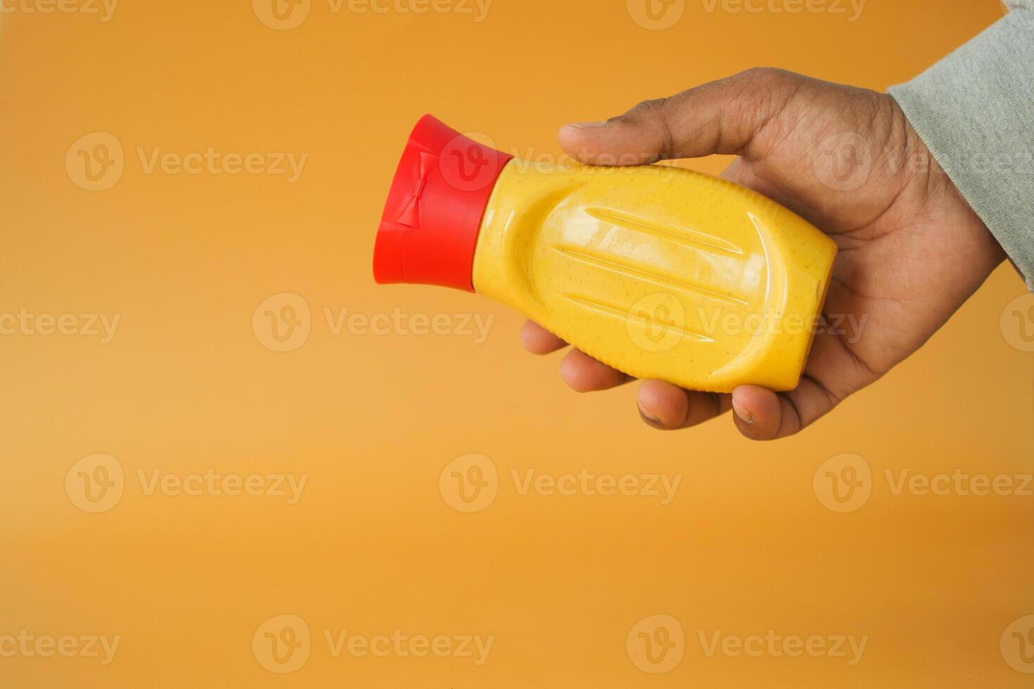 A hand gripping a bottle of mustard vividly set against a bright orange background photo