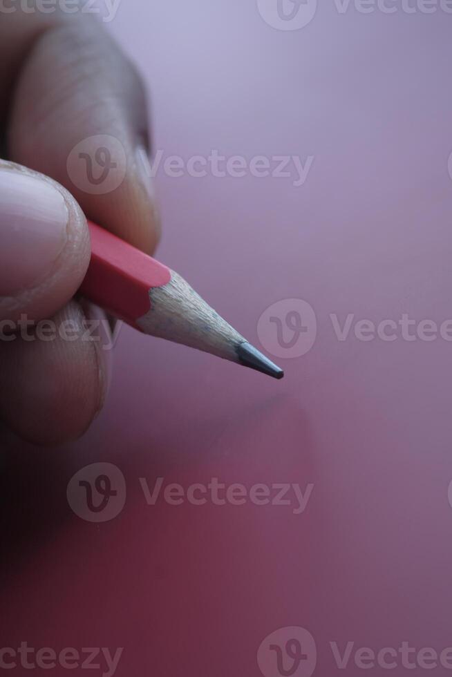 A closeup view of a hand firmly gripping a vibrant red pencil positioned on a soft pink surface photo