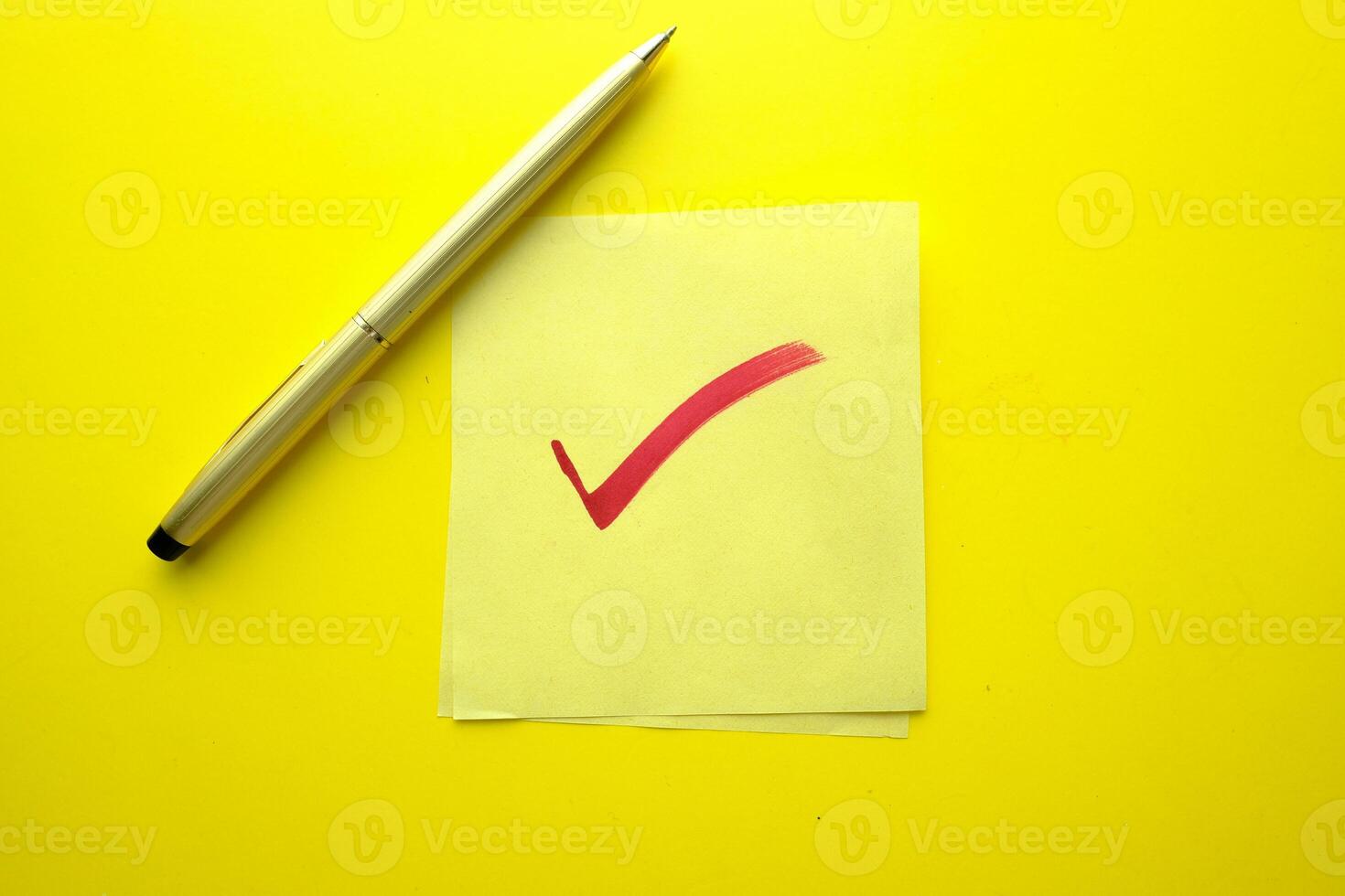 A Checkmark on a Sticky Note with a Pen resting beside it on a Bright Yellow Background photo