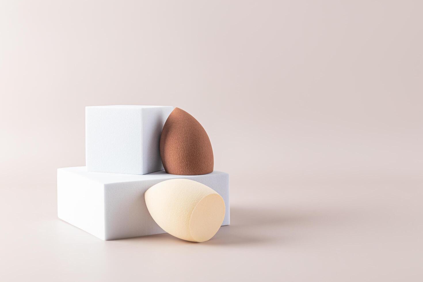 Beige and brown egg-shaped beauty sponge blenders on a square white catwalk and beige background. Front view. A sample of a makeup tool. A copy space photo