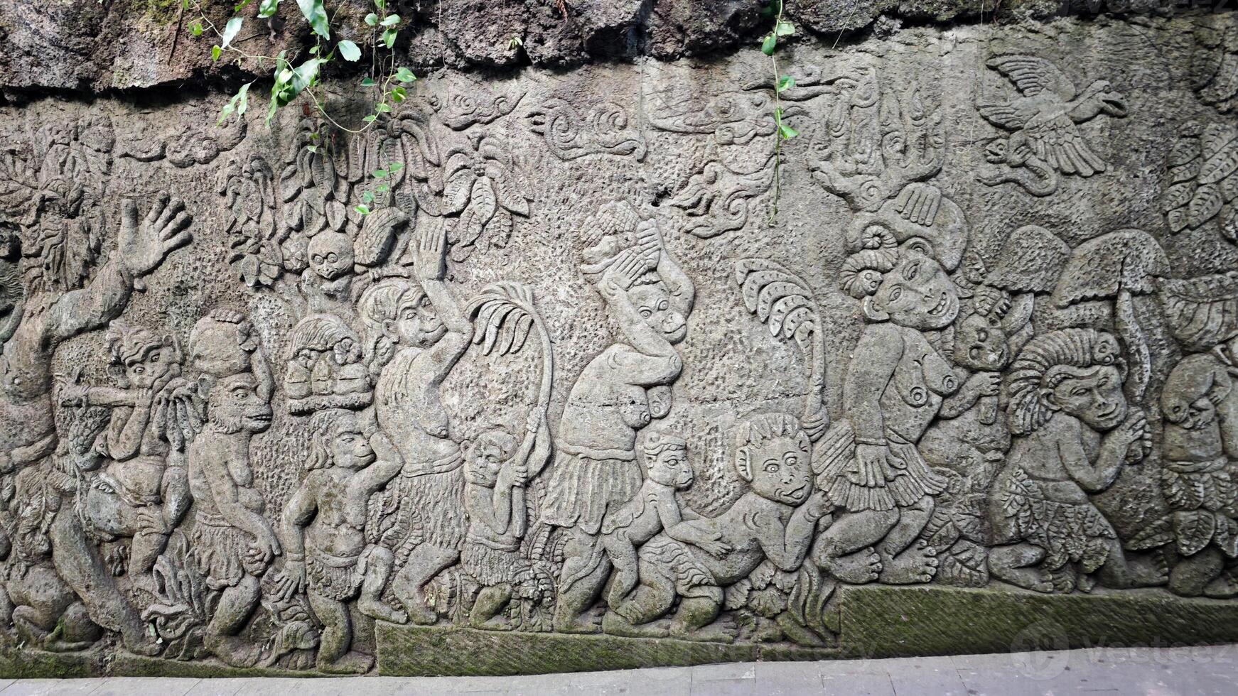 Intricately Carved Stone Wall Depicting Mythical Monkey Figures in a Lush Outdoor Setting Surrounded by Vines and Moss photo