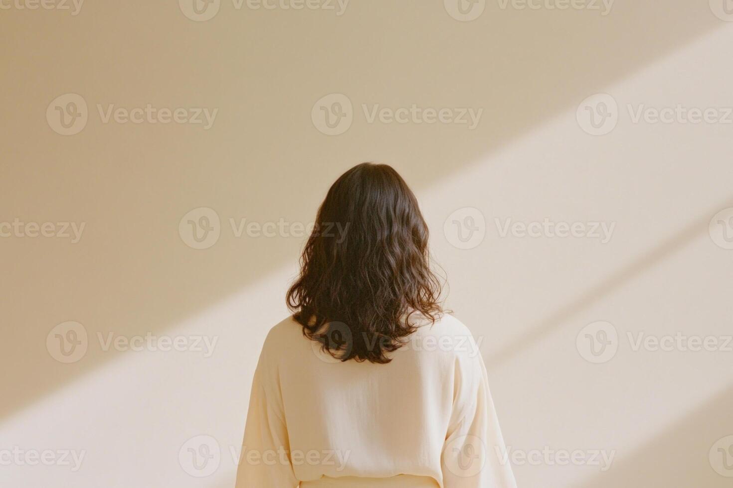 A woman in a stylish outfit stands gracefully against a pale taupe wall illuminated by soft natural light photo