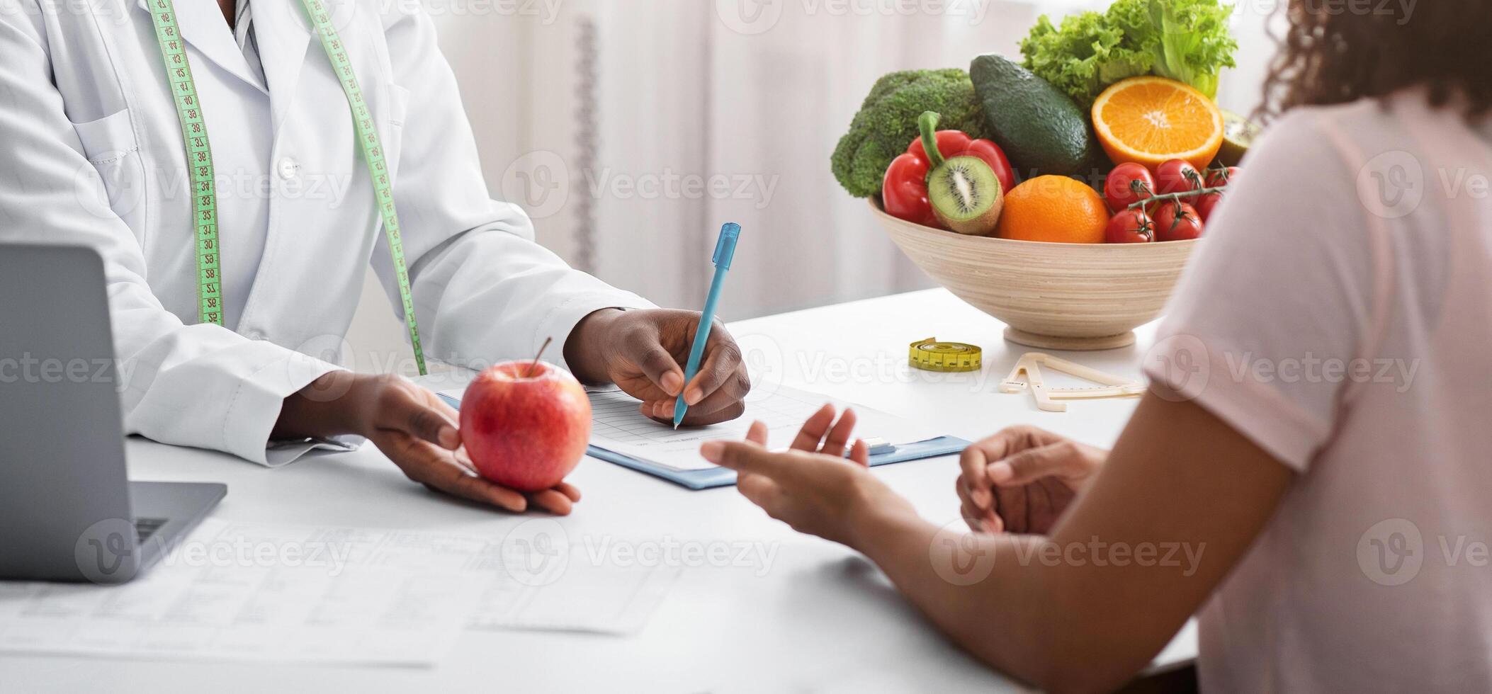 Dietician african woman giving young patient healthy diet tips, holding fresh red apple, clinic interior photo