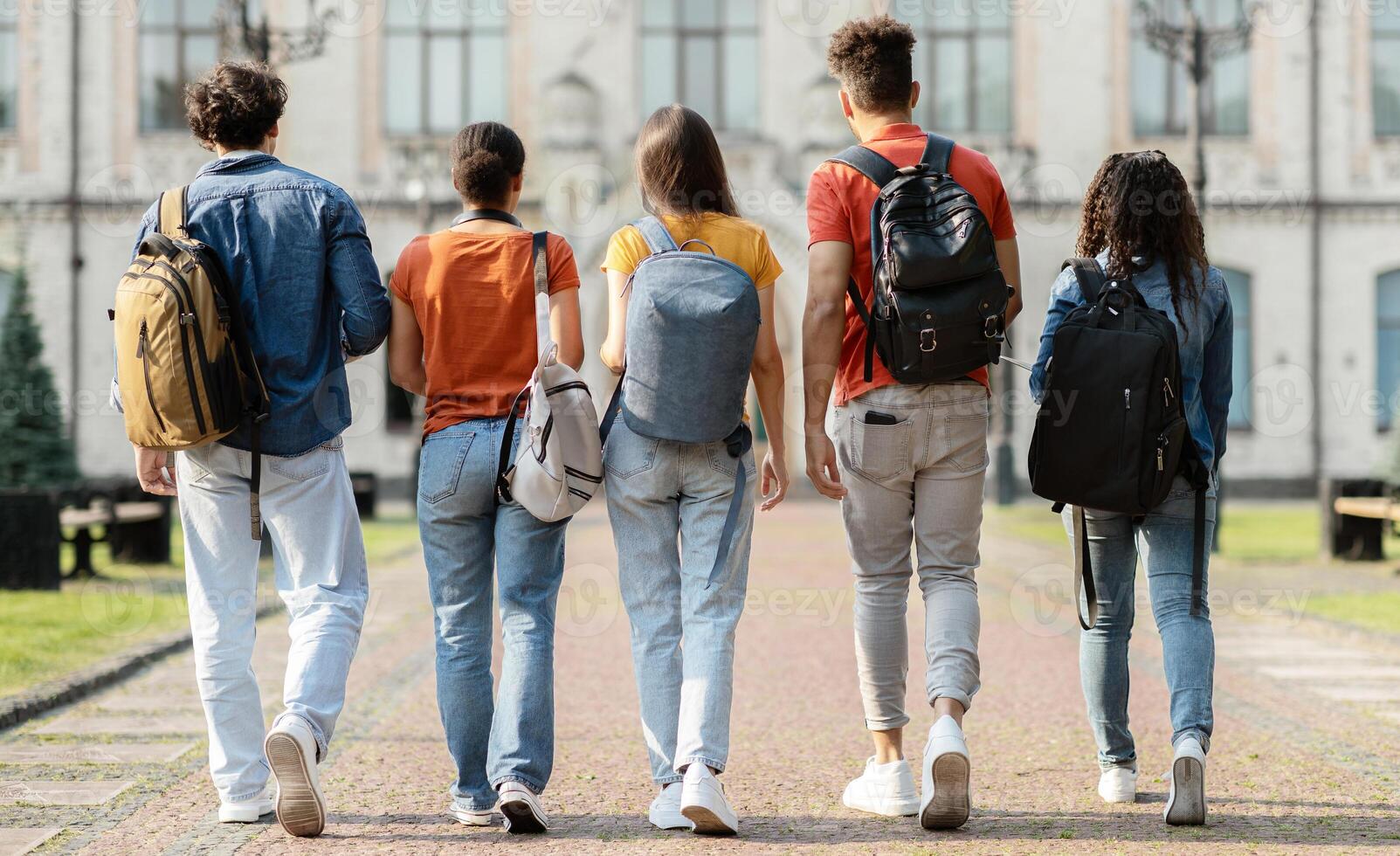Group of five students with backpacks walking at university campus together, rear view of multiethnic young people going to classes, diverse college friends spending time outdoors after lessons photo