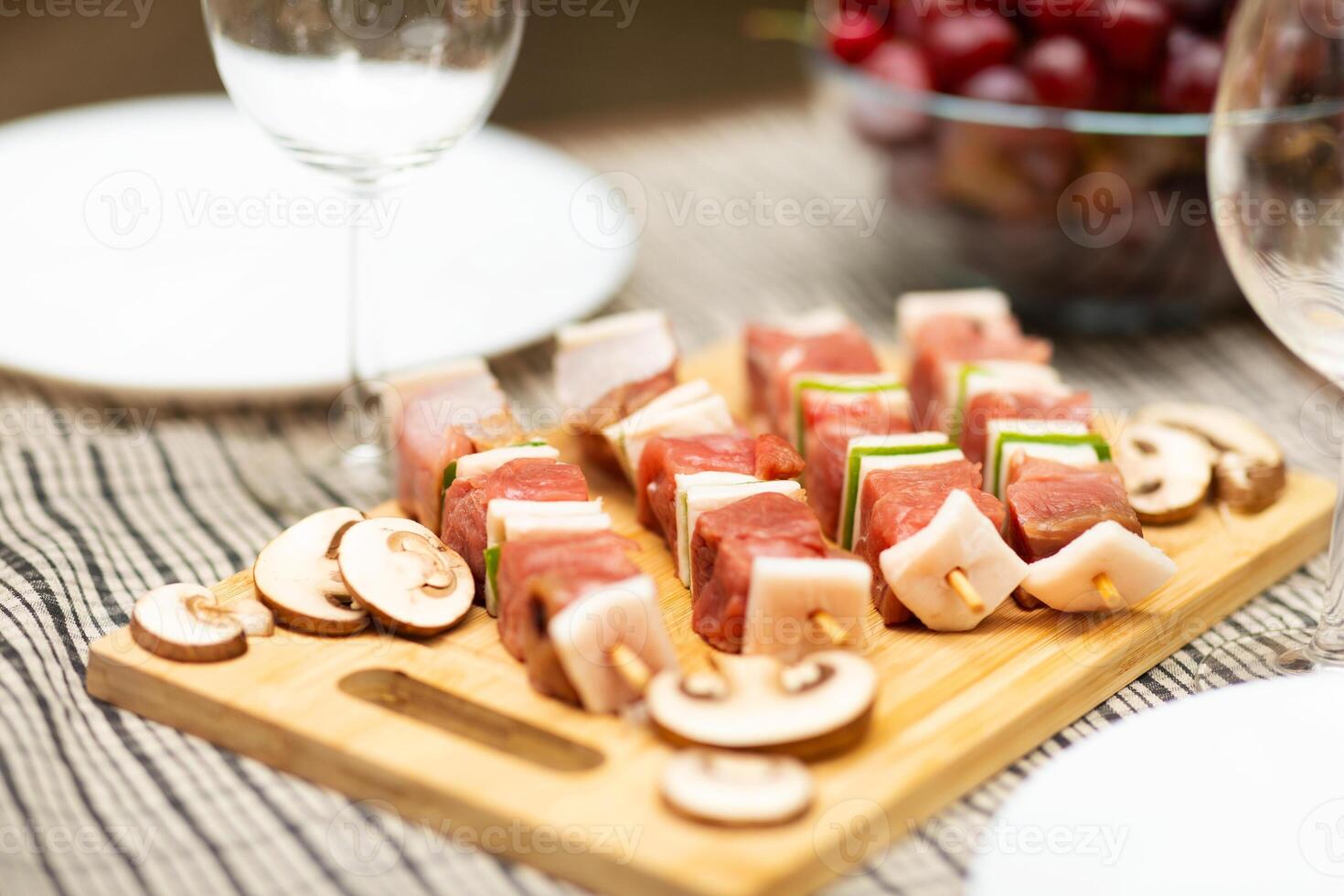 A wooden platter displays skewers of marinated meat and fresh vegetables, ready for grilling. Nearby, grapes rest in a bowl, along with empty wine glasses, hinting at an inviting gathering. photo