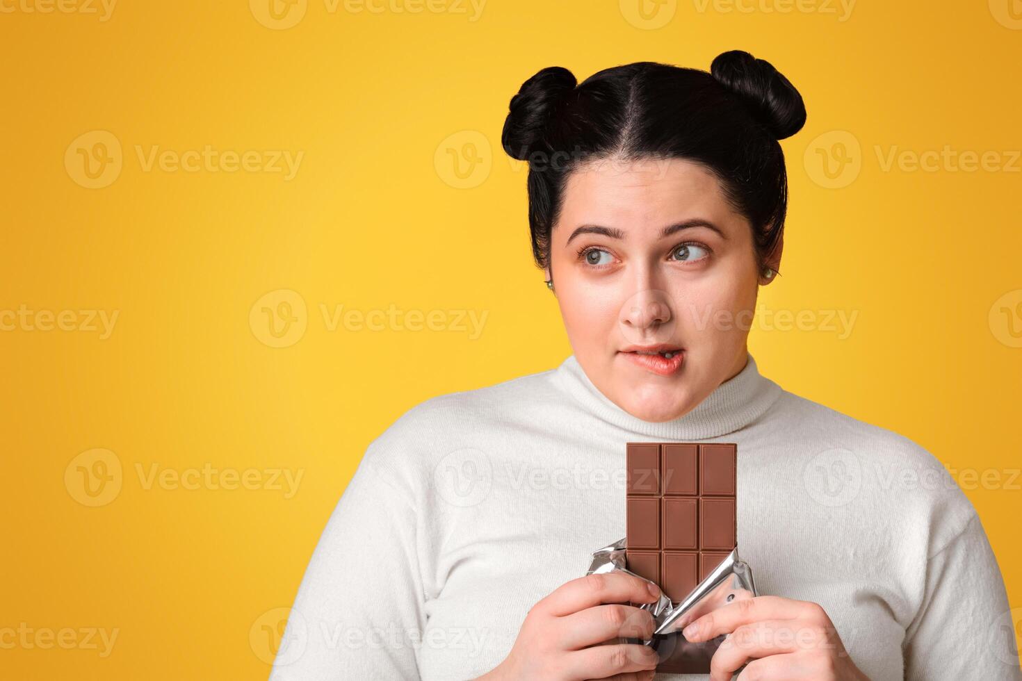 Sugar Addiction Concept. Young Obese Girl Cannot Resist Temptation To Eat Chocolate, Standing Over Yellow Background With Pensive Face Expression photo