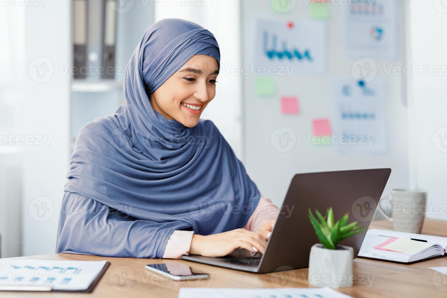 Office Job. Happy islamic woman in hijab working on laptop at desk, typing on keyboard and looking at computer screen. photo