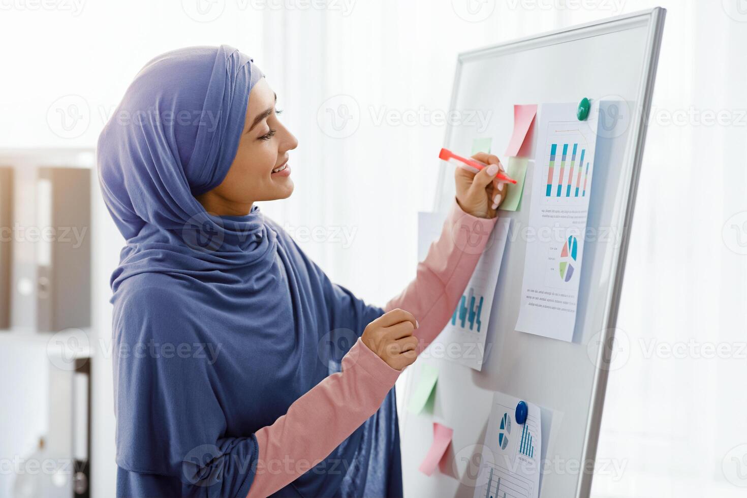 Islamic businesswoman in hijab preparing for presentation in office, taking notes, writing on white board with pinned graphs and charts photo