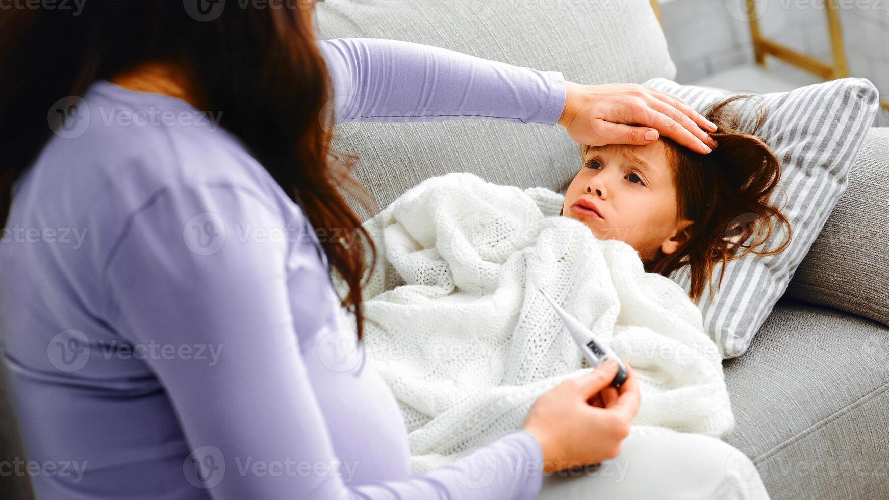 Sad little girl laying on sofa, feeling ill, complaining her pregnant mom, expecting mother checking her daughter's body temperature, close up photo