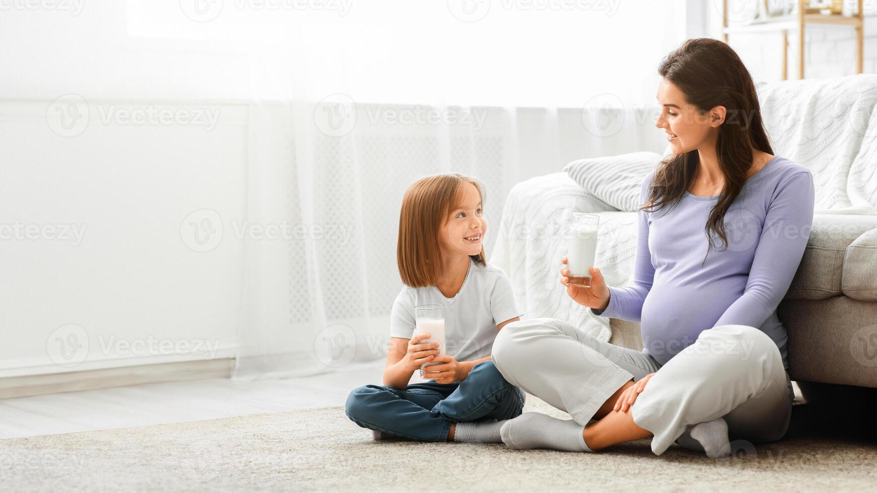 Healthy expecting young mom and little cute daughter having glass of milk, sitting on floor at living room photo