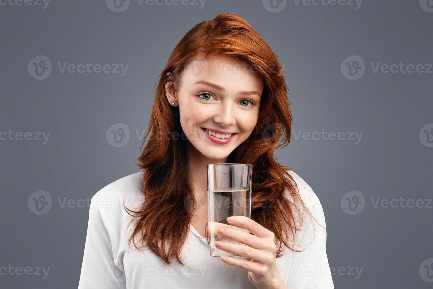 Positive Ginger-Haired Young Woman Holding Glass Of Water Smiling To Camera Standing Over White Studio Background. Youth, Healthy Lifestyle, Drink More Fresh Water Concept photo