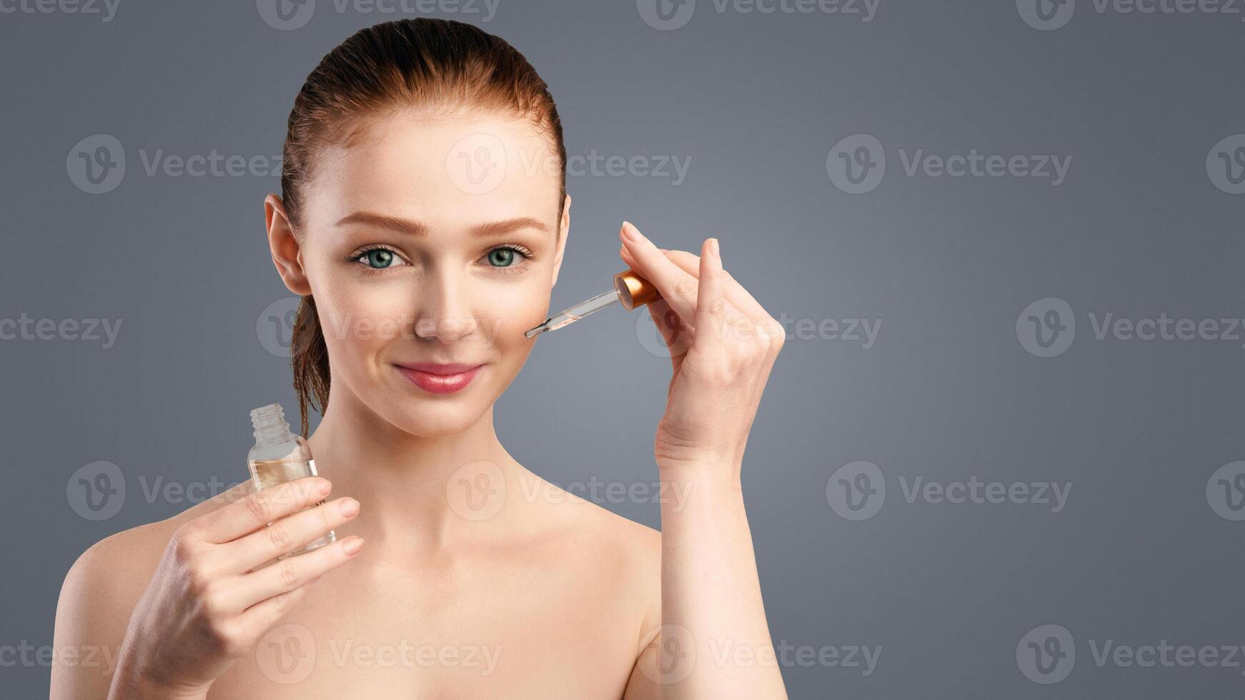 Skincare. Smiling Redhaired Woman Applying Cosmetic Oil On Face Caring For Facial Skin Posing Standing On White Background In Studio, Looking At Camera. Spa And Beauty Concept photo