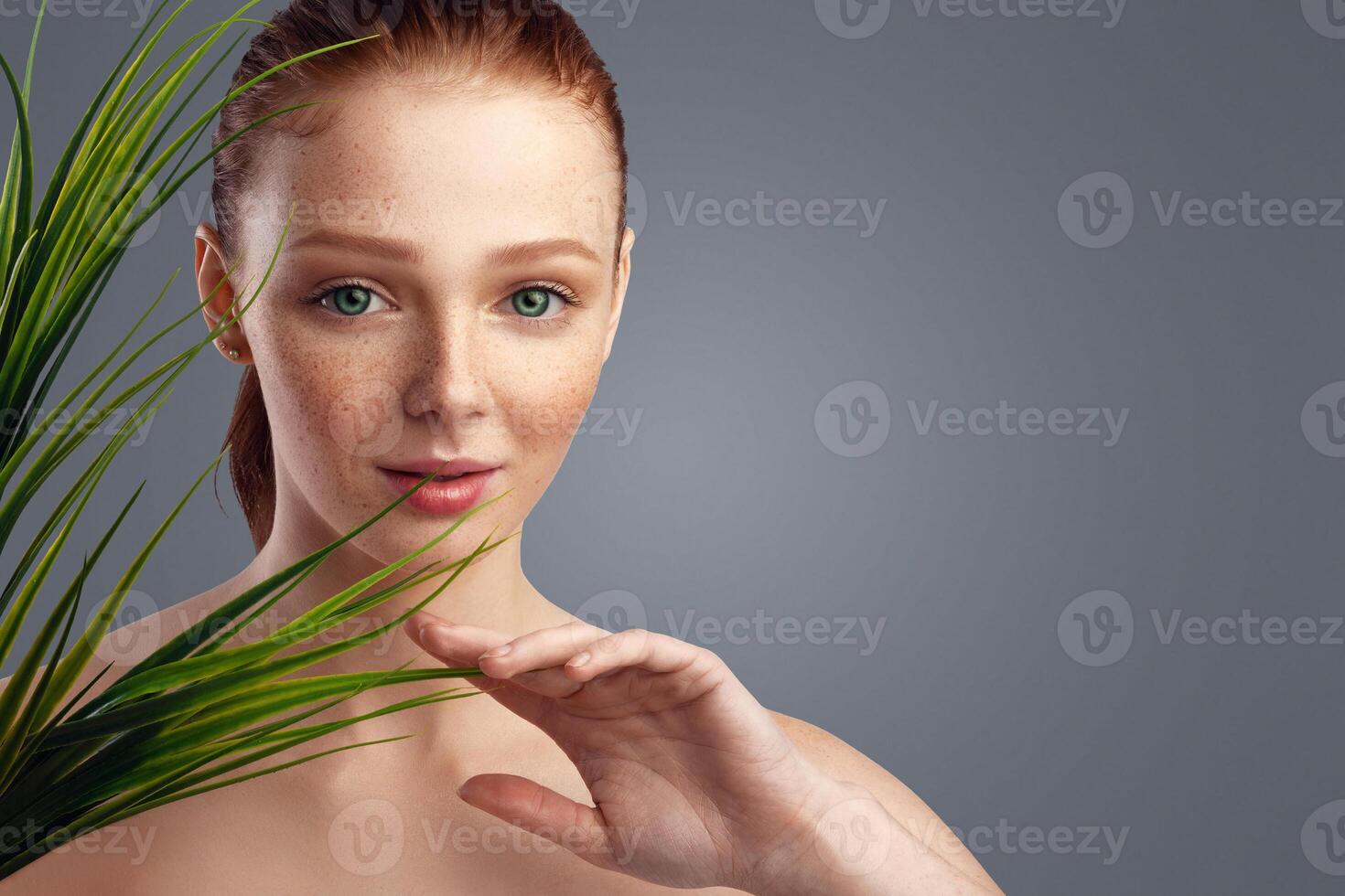 Red-Haired Millennial Female Touching Green Plant Posing Looking At Camera Standing Over Gray Background. Studio Portrait Of Young Lady Posing With Grass. Natural Beauty Care. Panorama, Empty Space photo