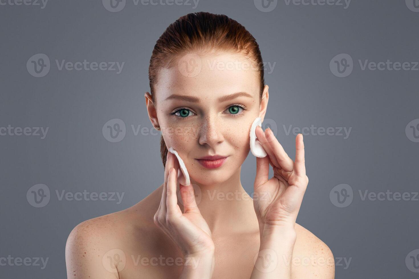 Red-Haired Young Woman Cleansing Skin Using Cotton Pads Posing On Gray Background In Studio, Looking At Camera. Natural Cosmetics For Facial Skincare Concept. Female Beauty Portrait photo