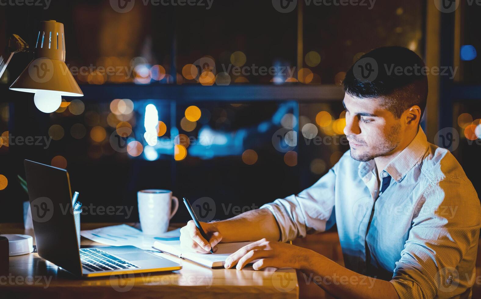 Business Quarantine. Freelancer Guy Taking Notes Sitting At Laptop Working Remotely Overnight In Home Office. Low Light, Free Space photo