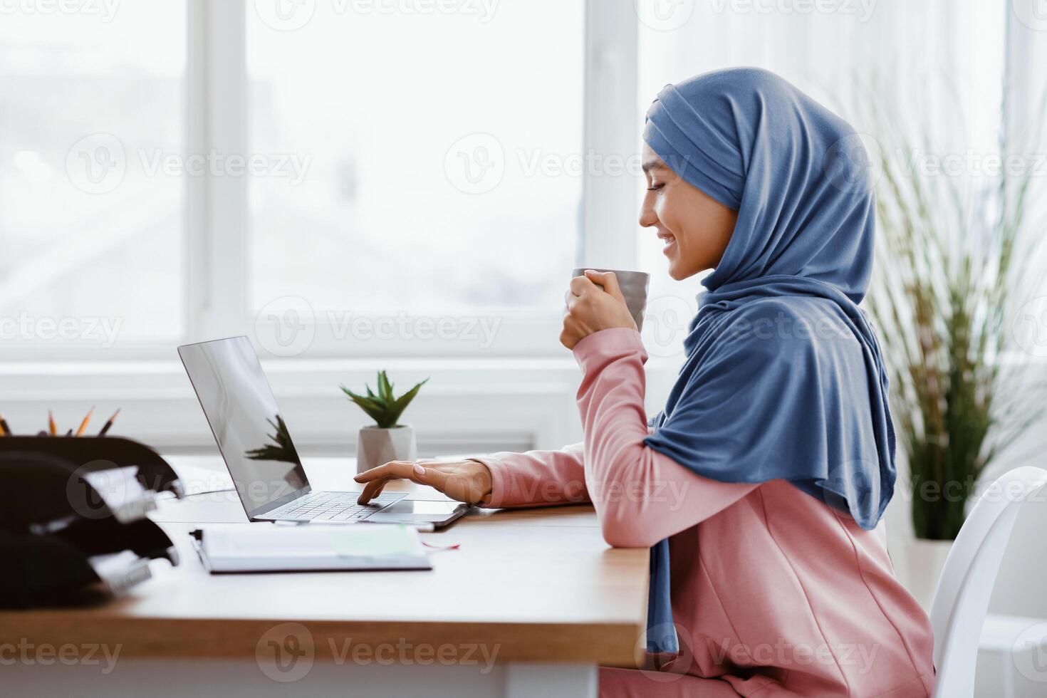 Young arabic businesswoman enjoying cup of coffee at her desk while working on laptop in modern office, side view with free space photo