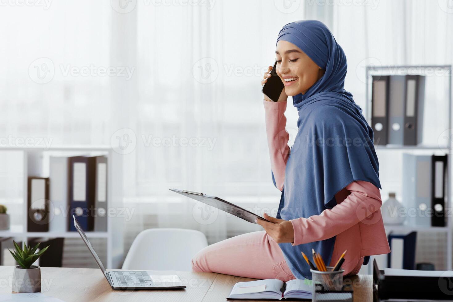 Portrait of happy arabic secretary talking on cellphone and holding clipboard, speaking with client, managing busines appointment photo