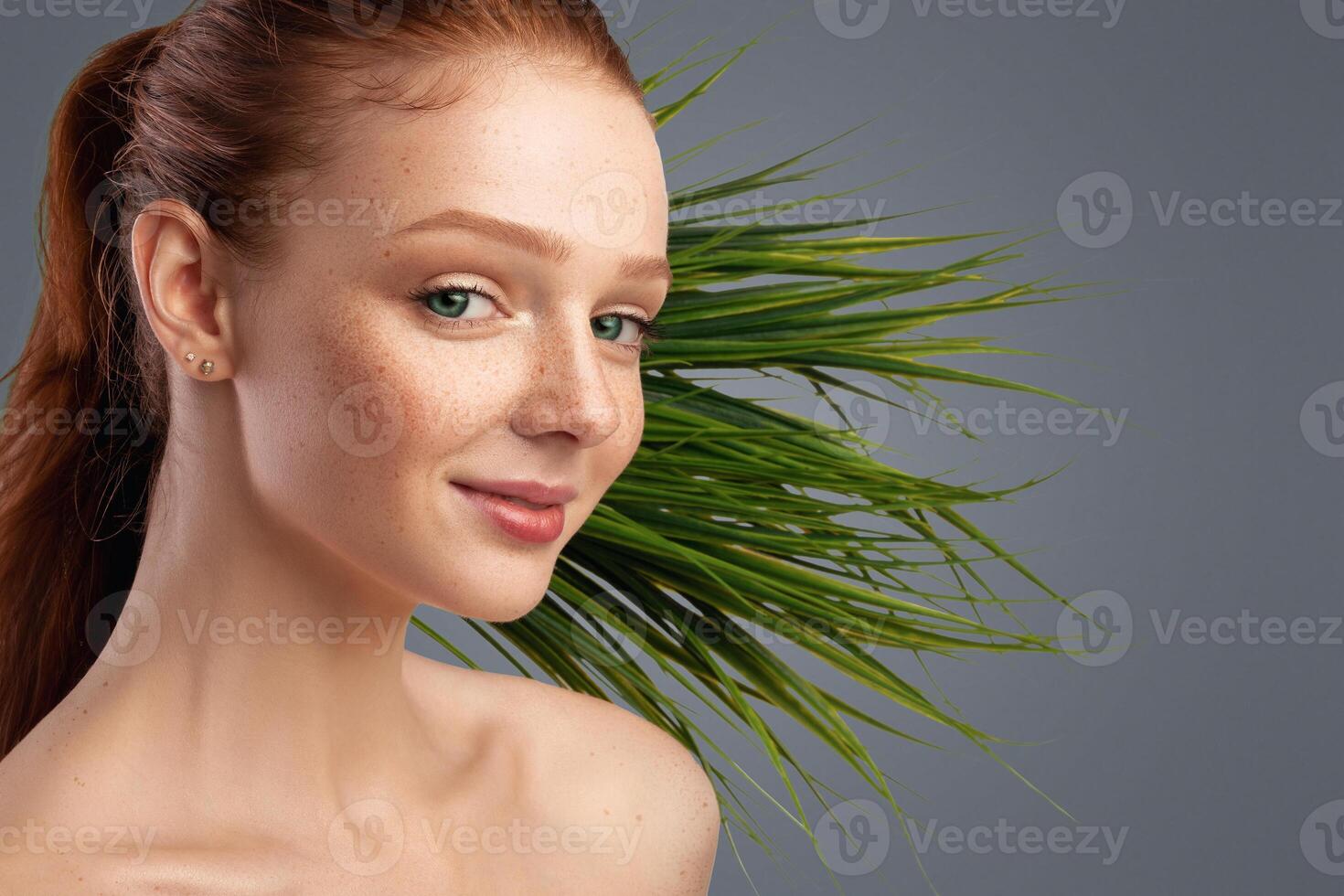Ginger-Haired Female Posing In Front Of Exotic Green Plant Smiling To Camera In Studio Over Gray Background. Panorama With Empty Space For Natural Cosmetics Advert photo