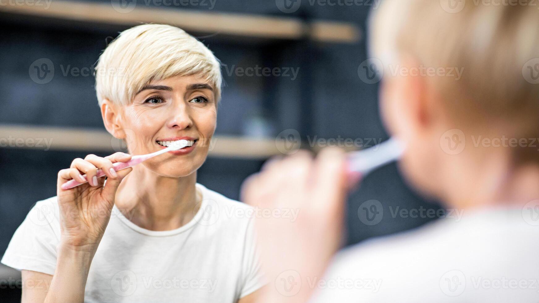 Toothcare Routine. Middle-Aged Woman Brushing Teeth Smiling To Reflection In Mirror Standing In Bathroom At Home. Selective Focus photo