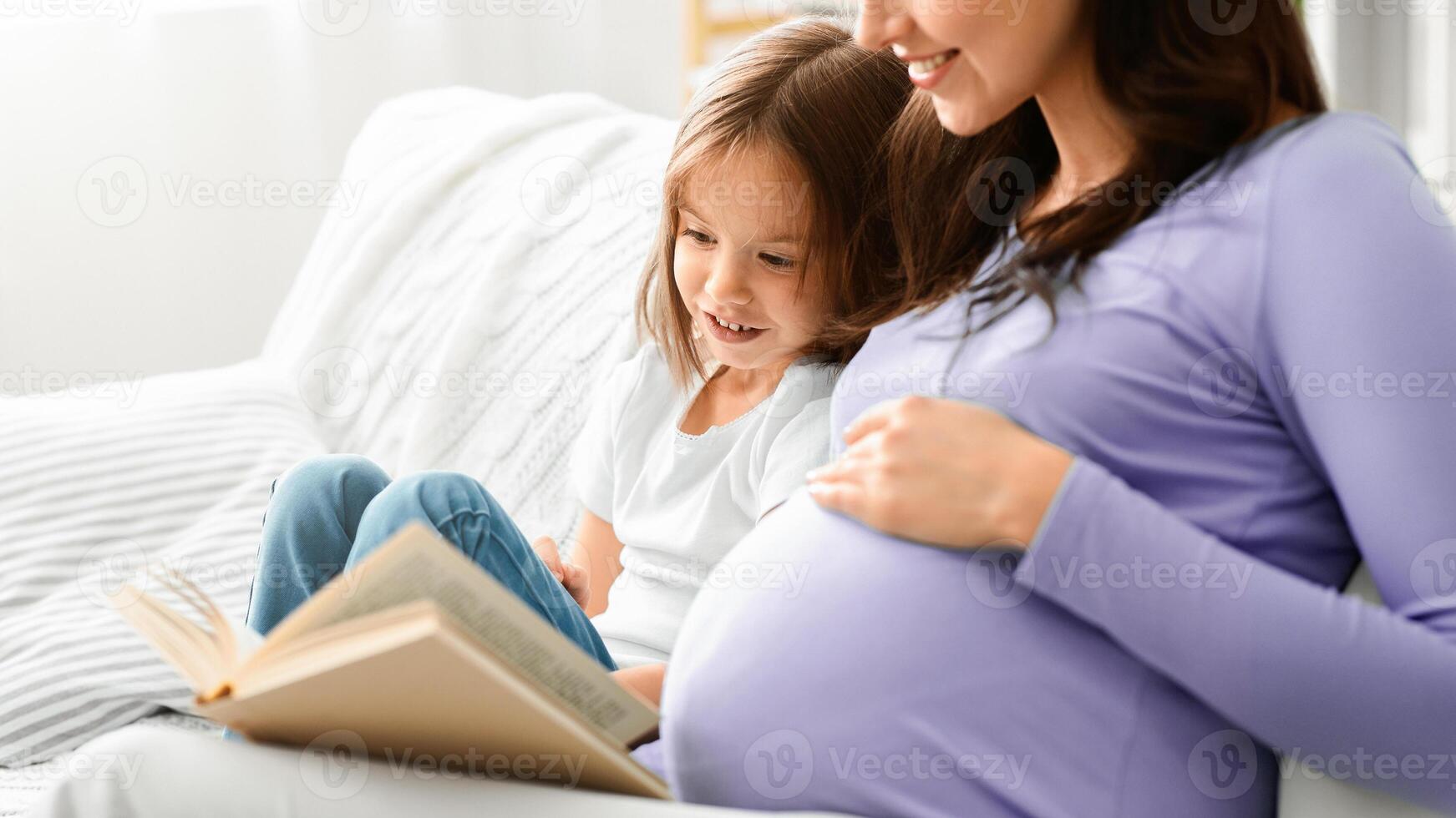 Close up of excited little girl reading book with expecting mom, home interior, copy space photo