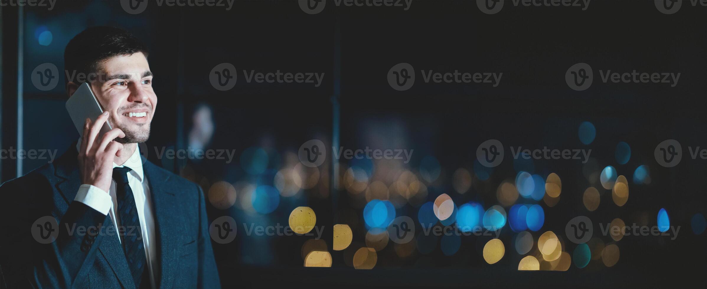 Smiling Entrepreneur Talking On Phone Standing Near Window With City View In Modern Office At Night. Business Communication. Copy Space photo