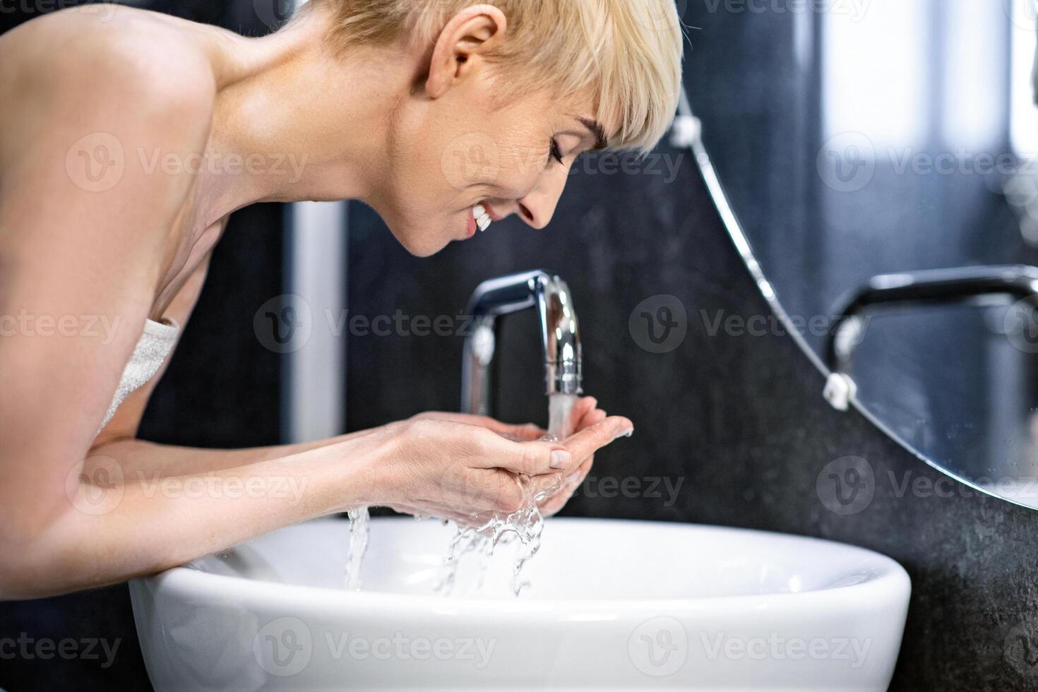 Joyful Mature Woman Washing Face Doing Morning Beauty Routine Standing Wrapped In Towel In Bathroom. Selective Focus photo