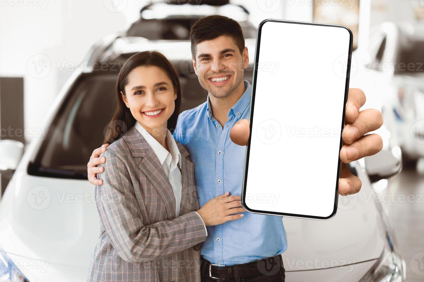 Ownership Concept. Young family embracing and standing together in front of auto in showroom, showing car keys at camera photo