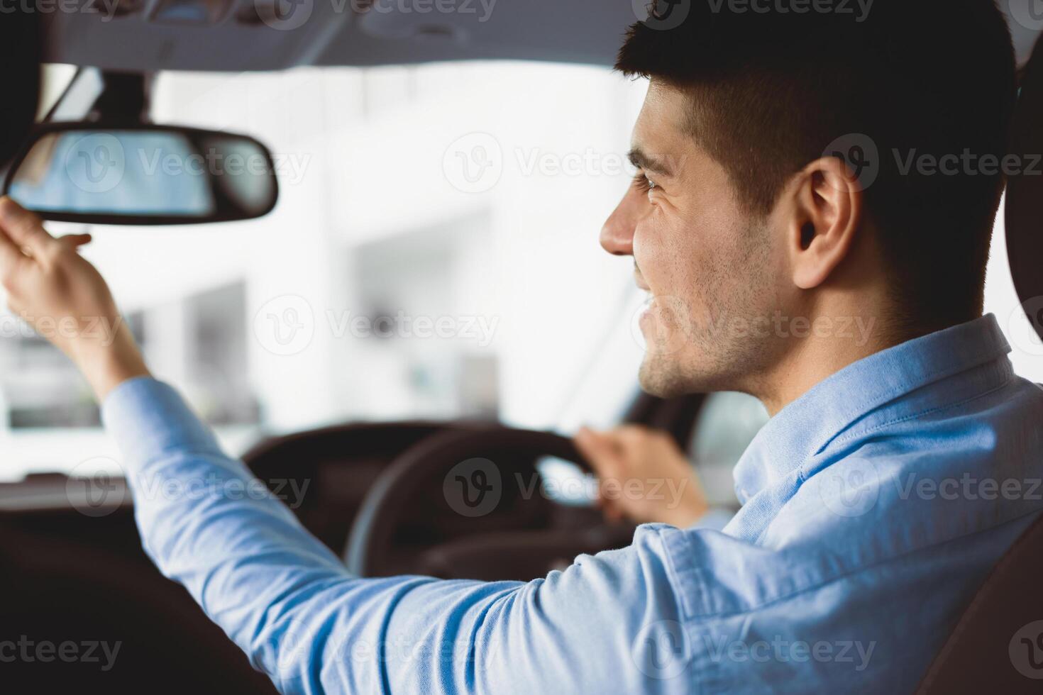 Close up back view portrait of smiling man in shirt adjusting rearview mirror while sitting in his car, selective focus photo